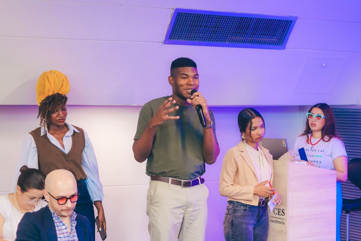 Joven habla con micrófono en un evento con otros participantes de pie